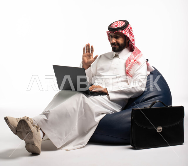 دردشة ومحادثات و تواصل اجتماعي ، التسوق الالكتروني وطلبات الاونلاين ، بورتريه لرجل عربي سعودي خليجي يرتدي الزي السعودي يجلس على كرسي مريح يستخدم الحاسوب المحمول، إنجاز مهام العمل عن بعد ، خلفية بيضاء