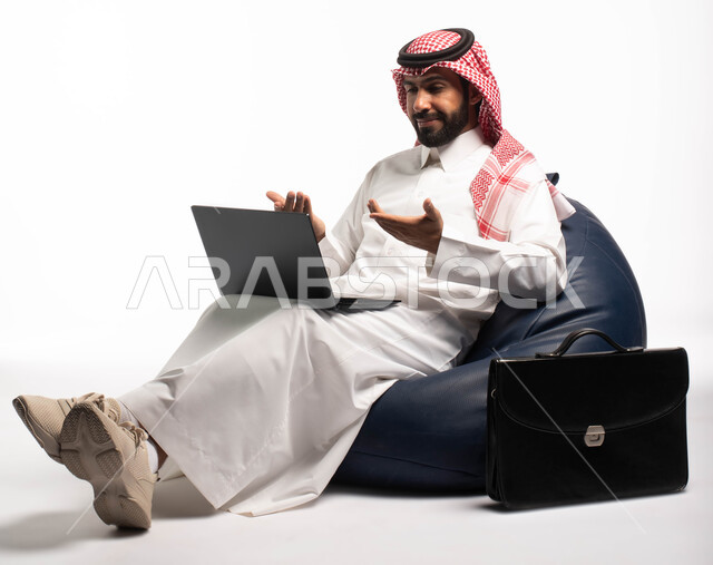 إيماءات وجه تدل على السعادة، دردشة ومحادثات و تواصل اجتماعي ، إنجاز مهام العمل عن بعد، التسوق الالكتروني وطلبات الاونلاين ، بورتريه لرجل عربي سعودي خليجي يرتدي الزي السعودي يجلس على كرسي مريح يستخدم الحاسوب المحمول، خلفية بيضاء