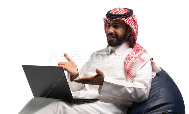 Working remotely using a laptop, live broadcasting and making a video call via a computer, a close-up portrait of a Saudi Arabian Gulf man wearing Saudi dress sitting on a comfortable chair, using a laptop, white background