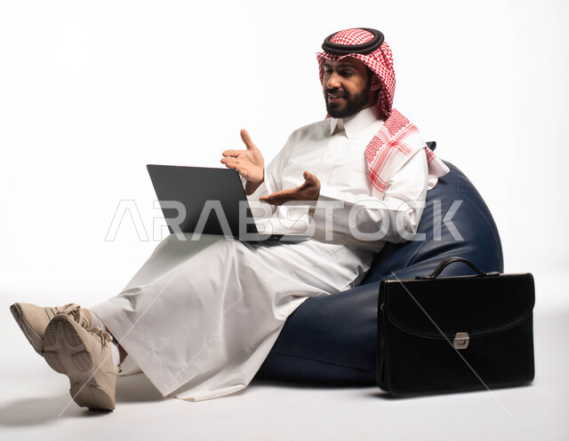 Live broadcast and making a video call via a computer, working remotely using a laptop, a portrait of a Saudi Arabian Gulf man wearing Saudi dress sitting on a comfortable chair next to a bag and using a laptop, white background