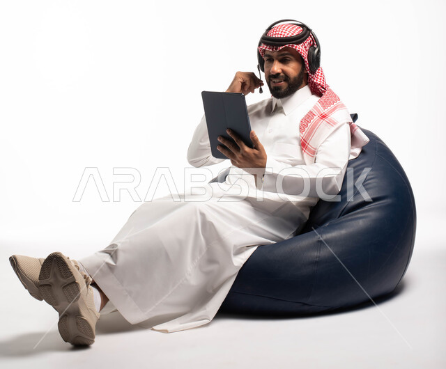 تسجيل الاغاني والصوتيات، تقديم البرامج الاذاعية،  بث مباشر واجراء مكالمة فيديو عبر الأجهزة الذكية ، بورتريه لشاب عربي سعودي خليجي يرتدي الزي التقليدي يجلس على كرسي مريح، رجل يضع سماعة الرأس ويستخدم جهاز التابلت ، خلفية بيضاء