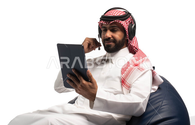 Presenting radio programs, live broadcasting and making a video call via smart devices, a close-up portrait of a Saudi Arabian Gulf youth wearing traditional dress sitting on a comfortable chair, a man wearing a headphone and using a tablet, recording son