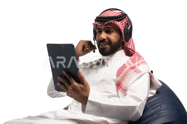 تسجيل الاغاني والصوتيات، تقديم البرامج الاذاعية،  بث مباشر واجراء مكالمة فيديو عبر الأجهزة الذكية ، بورتريه مقرب لشاب عربي سعودي خليجي يرتدي الزي التقليدي يجلس على كرسي مريح، رجل يضع سماعة الرأس ويستخدم جهاز التابلت ، خلفية بيضاء