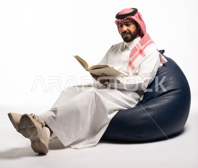 قضاء الوقت بالقراءة والتعلم والثقافة، التمعن والتركيز، بورتريه لرجل عربي سعودي خليجي يرتدي الزي السعودي التقليدي يجلس على كرسي مريح ويحمل بيديه كتاب، خلفية بيضاء