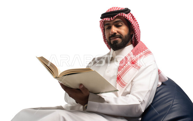 قضاء الوقت بالقراءة والتعلم والثقافة، التمعن والتركيز، بورتريه مقرب لرجل عربي سعودي خليجي يرتدي الزي السعودي التقليدي يجلس على كرسي مريح ويحمل بيديه كتاب، خلفية بيضاء