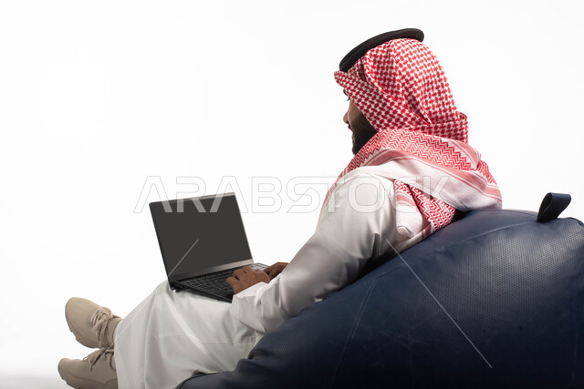  دردشة ومحادثات و تواصل اجتماعي ، إنجاز مهام العمل عن بعد، التسوق الالكتروني وطلبات الاونلاين ، بورتريه من الخلف لرجل عربي سعودي خليجي يرتدي الزي السعودي يجلس على كرسي مريح يستخدم الحاسوب المحمول، خلفية بيضاء