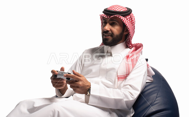 قضاء وقت ممتع، التفاعل والاندماج مع ألعاب البلايستيشن، تقنيات حديثة مسلية, بورتريه لرجل عربي سعودي خليجي يرتدي الزي التقليدي يجلس على كرسي مريح ويحمل بيده عصا التحكم  ويلعب بألعاب الفيديو ، خلفية بيضاء