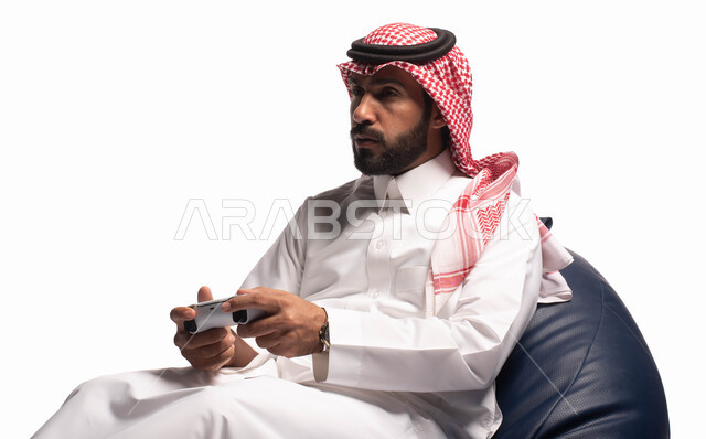 Interaction and integration with PlayStation games, modern entertaining technologies, a portrait of a Saudi Arabian Gulf man dressed in traditional dress, sitting on a comfortable chair, holding a joystick in his hand, and playing video games, white backg