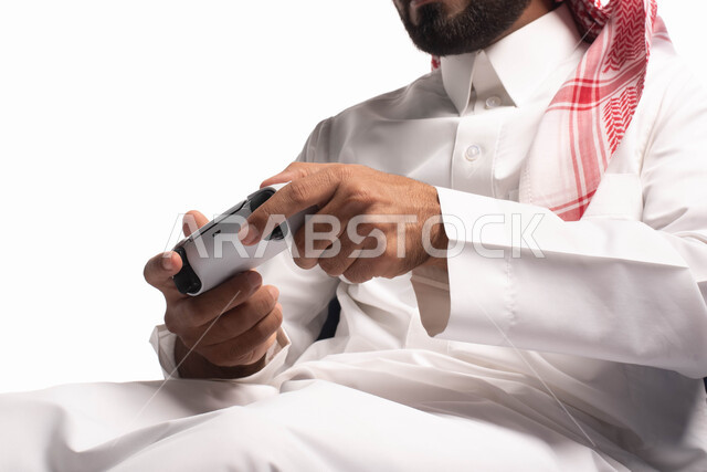 التفاعل والاندماج مع ألعاب البلايستيشن، تقنيات حديثة مسلية, بورتريه لرجل عربي سعودي خليجي يرتدي الزي التقليدي يجلس على كرسي مريح ويحمل بيده عصا التحكم  ويلعب بألعاب الفيديو ، خلفية بيضاء