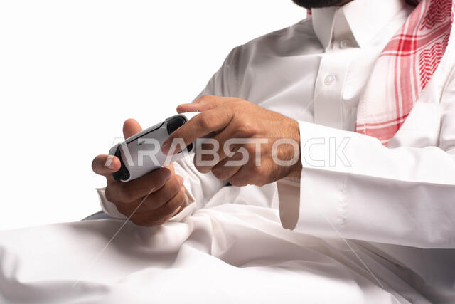 تقنيات حديثة مسلية, قضاء وقت ممتع ومسلي، التفاعل والاندماج مع ألعاب البلايستيشن، بورتريه مقرب لرجل عربي سعودي خليجي يرتدي الزي التقليدي يجلس على كرسي مريح ويحمل بيده عصا التحكم  ويلعب بألعاب الفيديو ، خلفية بيضاء