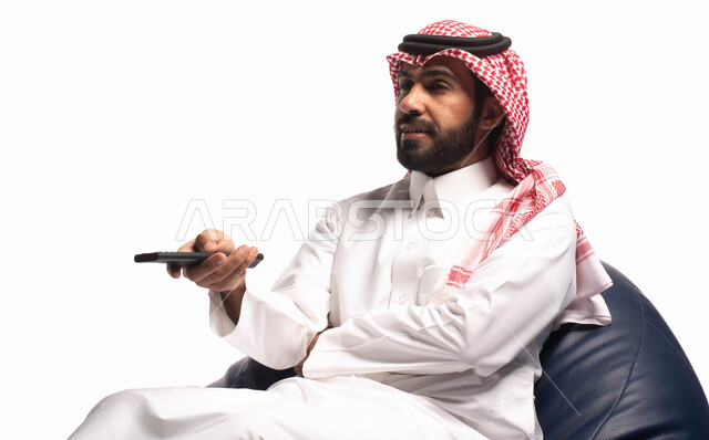 استخدام عصا التحكم, قضاء أوقات ممتعة ومسلية، ايماءات وجه تدل على السعاده والاستمتاع, بورتريه مقرب لرجل عربي سعودي خليجي يجلس على كرسي مريح ويشاهد التلفاز, التحكم بالتلفاز عن بعد, خلفية بيضاء. 