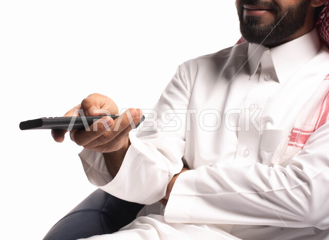 قضاء أوقات ممتعة ومسلية، ايماءات وجه تدل على السعاده والاستمتاع, بورتريه مقرب لرجل عربي سعودي خليجي يجلس على كرسي مريح ويشاهد التلفاز, استخدام عصا التحكم, التحكم بالتلفاز عن بعد, خلفية بيضاء. 