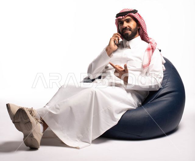 إتصال و تعارف وعلاقات اجتماعية ودردشة ومحادثات و تواصل اجتماعي، إجراء مكالمة هاتفية ،بورتريه لرجل عربي سعودي خليجي مبتسم يرتدي الزي التقليدي يجلس على كرسي مريح بيده هاتف محمول ، خلفية بيضاء