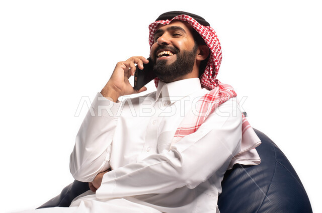 إتصال و تعارف وعلاقات اجتماعية ودردشة ومحادثات و تواصل اجتماعي، إجراء مكالمة هاتفية ،بورتريه مقرب لرجل عربي سعودي خليجي مبتسم يرتدي الزي السعودي يجلس على كرسي مريح بيده هاتف محمول ، خلفية بيضاء
