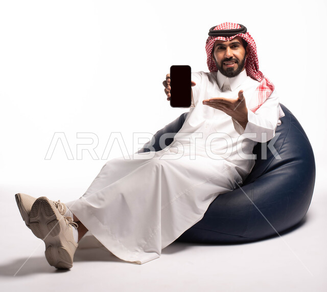 عرض شاشة سوداء فارغة ، بورتريه لرجل عربي خليجي سعودي يرتدي الزي التقليدي بجلس على كرسي مريح ويحمل بيده الهاتف المحمول ، النظر إلى الكاميرا ، ايماءات وجه تدل على الفرح والسرور ، الاهتمام بالمظهر الخارجي ،  خلفية بيضاء