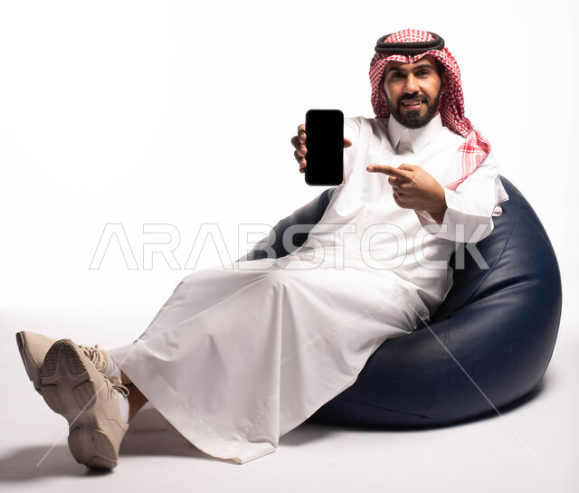 عرض شاشة سوداء فارغة ، بورتريه لرجل عربي خليجي سعودي يرتدي الزي التقليدي بجلس على كرسي مريح ويحمل بيده الهاتف المحمول ، النظر إلى الكاميرا ، ايماءات وجه تدل على الفرح والسرور ، الاهتمام بالمظهر الخارجي ،  خلفية بيضاء