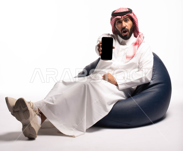 عرض شاشة سوداء فارغة ، بورتريه لرجل عربي خليجي سعودي يرتدي الزي التقليدي بجلس على كرسي مريح ويحمل بيده الهاتف المحمول ، النظر إلى الكاميرا ، ايماءات وجه تدل على الدهشة، الاهتمام بالمظهر الخارجي ،  خلفية بيضاء