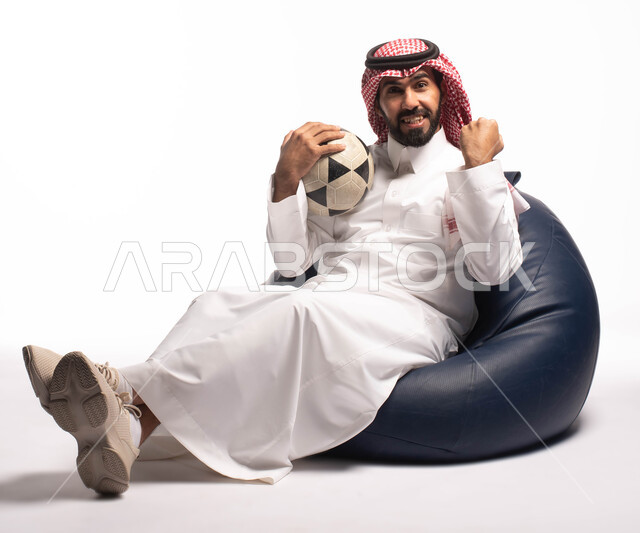 الفرح بالفوز والإنتصار, بورتريه لرجل عربي سعودي خليجي يرتدي الزي التقليدي يجلس على كرسي يرفع يديه مضموم الكفين يقوم بالتشجيع, إيماءات وجه ويدين تدل على السعاده, التفاعل والإندماج مع المباريات, أنديه وتشجيع,  خلفية بيضاء