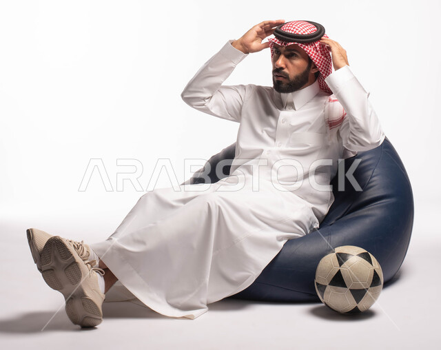 التفاعل والاندماج مع المباريات، أندية وتشجيع، بورتريه لرجل عربي سعودي خليجي يرتدي الزي التقليدي يجلس على كرسي مريح يضع يديه على رأسه, إيماءات وجه ويدين تدل على الحزن والخساره وعدم الفوز بالمباراه, خلفية بيضاء