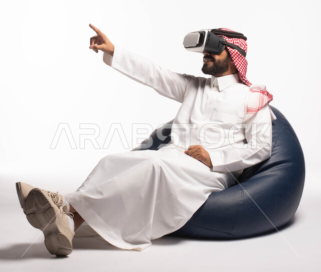 الاستمتاع بمشاهدة أفلام الواقع الافتراضي، استخدام حاسة اللمس لتحريك العناصر الهولوجرامية، بورتريه لرجل عربي خليجي سعودي يجلس على كرسي مريح ويرتدي نظارة الواقع الافتراضي ثلاثية الابعاد، خلفية بيضاء
