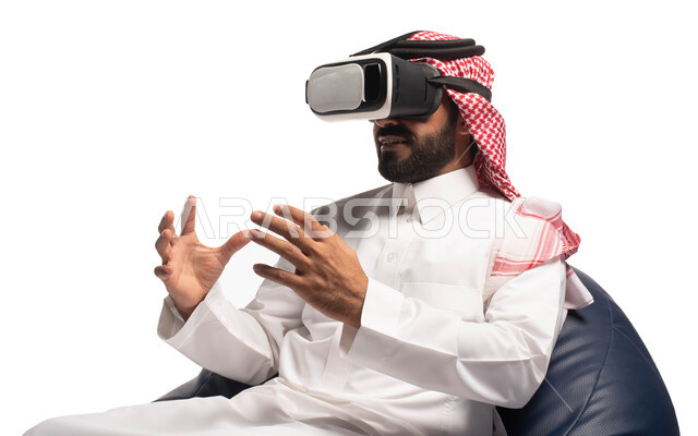 الاستمتاع بمشاهدة أفلام الواقع الافتراضي، استخدام حاسة اللمس لتحريك العناصر الهولوجرامية، بورتريه مقرب لرجل عربي خليجي سعودي يجلس على كرسي مريح ويرتدي نظارة الواقع الافتراضي ثلاثية الابعاد، خلفية بيضاء