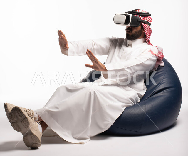 الاستمتاع بمشاهدة أفلام الواقع الافتراضي، استخدام حاسة اللمس لتحريك العناصر الهولوجرامية، بورتريه لرجل عربي خليجي سعودي يجلس على كرسي مريح ويرتدي نظارة الواقع الافتراضي ثلاثية الابعاد، خلفية بيضاء