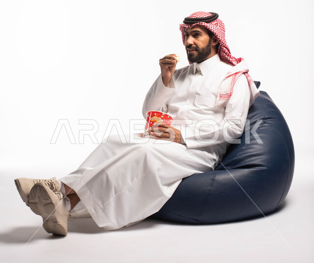 قضاء أوقات ممتعة ومسلية، بورتريه رجل عربي خليجي سعودي يرتدي الزي التقليدي يحمل بيده علبة الفوشار ومستمتع بمشاهدة الأفلام ، إيماءات وجه ويدين تدل على السعادة والمتعة، مشاهدة فلم سينمائي، التسليه والترفيه، خلفية بيضاء