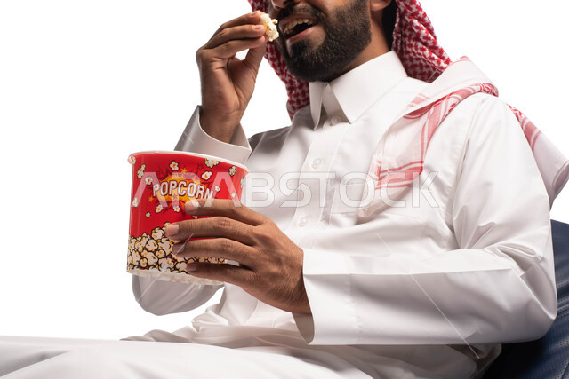 Watching a movie, spending fun and entertaining times, a close-up portrait of a Saudi Arabian Gulf man wearing traditional dress holding a box of popcorn in his hand and enjoying watching movies, face and hand gestures indicating happiness and enjoyment, 