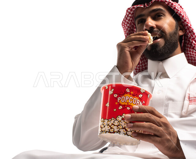 قضاء أوقات ممتعة ومسلية، مشاهدة فلم سينمائي، بورتريه مقرب لرجل عربي خليجي سعودي يرتدي الزي التقليدي يحمل بيده علبة الفوشار ومستمتع بمشاهدة الأفلام ، إيماءات وجه ويدين تدل على السعادة والمتعة، خلفية بيضاء