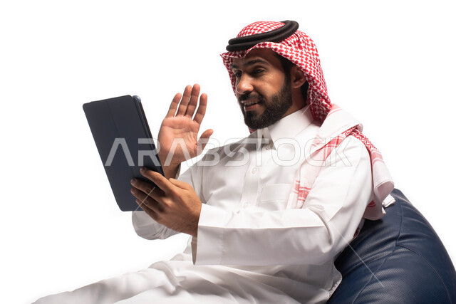 إجراء مكالمات الفيديو عبر الانترنت، استخدام اجهزة تقنية حديثة، بورتريه  مقربة لرجل عربي سعودي خليجي يجلس على كرسي مريح يستخدم جهاز التابلت، تصفح الإنترنت وإنجاز المهام عن بعد ، الشراء والبيع عبر الاونلاين، خلفية بيضاء 