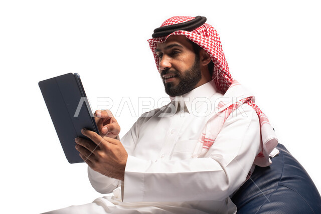 Working and completing tasks remotely, making video calls over the Internet, using modern technology devices, a close-up portrait of a Saudi Arabian Gulf man wearing traditional dress, sitting on a comfortable chair and using a tablet, white background