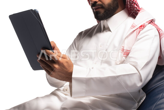Using modern technical devices, working and completing tasks remotely, making video calls over the Internet, a close-up portrait of a Saudi Arabian Gulf man wearing traditional dress, sitting on a comfortable chair and using a tablet, white background