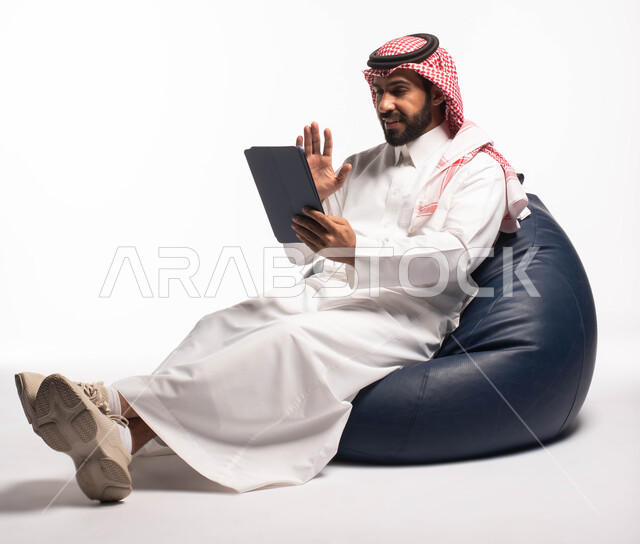  تصفح الإنترنت وإنجاز المهام عن بعد، إجراء مكالمات الفيديو عبر النت، استخدام اجهزة تقنية حديثة، بورتريه لرجل عربي سعودي خليجي يجلس على كرسي مريح يستخدم جهاز التابلت، الشراء والبيع عبر الاونلاين، خلفية بيضاء 