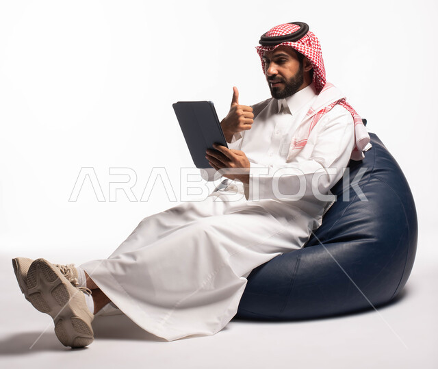 قضاء وقت ممتع، إنجاز المهام عن بعد، إجراء مكالمات الفيديو عبر الانترنت، استخدام اجهزة تقنية حديثة، بورتريه لرجل عربي سعودي خليجي يرتدي الزي التقليدي يجلس على كرسي مريح ويستخدم جهاز التابلت، خلفية بيضاء 