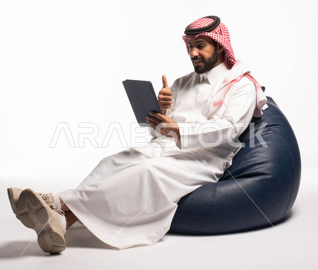  تصفح برامج التواصل الاجتماعي  وإنجاز المهام عن بعد، إجراء مكالمات الفيديو عبر الانترنت، استخدام اجهزة تقنية حديثة، بورتريه لرجل عربي سعودي خليجي يرتدي الزي التقليدي يجلس على كرسي مريح ويستخدم جهاز التابلت، خلفية بيضاء 