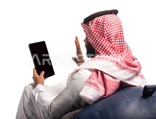 Making video calls over the Internet, using modern technology devices, a close-up portrait of a Saudi Arabian Gulf man sitting on a comfortable chair using a tablet, surfing the Internet and completing tasks remotely, buying and selling via the Internet, 