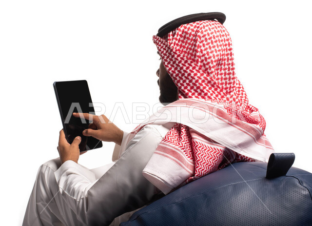 استخدام اجهزة تقنية حديثة، بورتريه  مقربة من الخلف لرجل عربي سعودي خليجي يجلس على كرسي مريح يستخدم جهاز التابلت ، التسوق الإلكتروني الآمن، تصفح الإنترنت وإنجاز المهام عن بعد ، الشراء والبيع عبر الاونلاين، خلفية بيضاء 