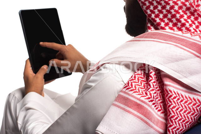 استخدام اجهزة تقنية حديثة، بورتريه  مقربة من الخلف لرجل عربي سعودي خليجي يجلس على كرسي مريح يستخدم جهاز التابلت ، التسوق الإلكتروني الآمن، تصفح الإنترنت وإنجاز المهام عن بعد ، الشراء والبيع عبر الاونلاين، خلفية بيضاء 