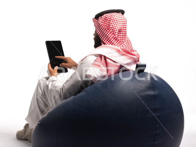 إجراء مكالمات الفيديو عبر الانترنت، استخدام اجهزة تقنية حديثة، بورتريه  مقربة من الخلف لرجل عربي سعودي خليجي يجلس على كرسي مريح يستخدم جهاز التابلت، تصفح الإنترنت وإنجاز المهام عن بعد ، الشراء والبيع عبر الاونلاين، خلفية بيضاء 