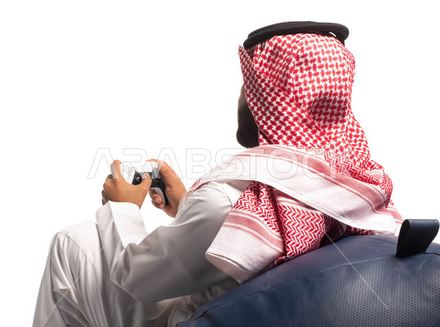 قضاء وقت الفراغ باللعب بألعاب البلايستيشن، تقنيات حديثة مسلية, بورتريه من الخلف لرجل عربي سعودي خليجي يرتدي الزي التقليدي يجلس على كرسي مريح ويحمل بيده عصا التحكم  ويلعب بألعاب الفيديو ، خلفية بيضاء