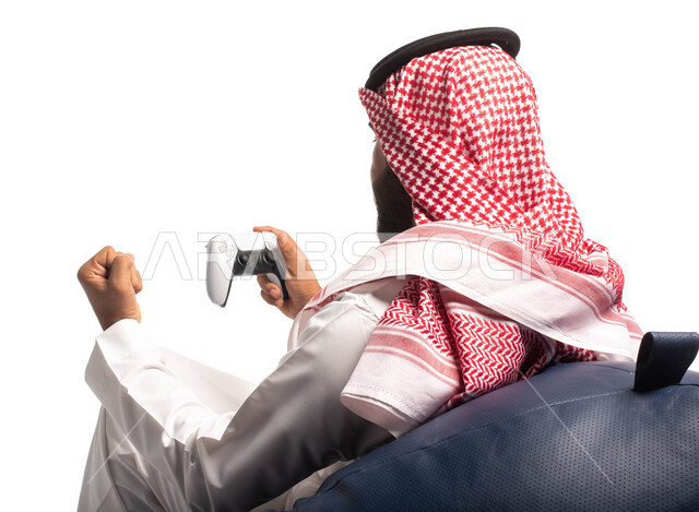 ايماءات يدين تدل على الفوز والانتصار بألعاب البلايستيشن، تقنيات حديثة مسلية, بورتريه من الخلف لرجل عربي سعودي خليجي يرتدي الزي التقليدي يجلس على كرسي مريح ويحمل بيده عصا التحكم  ويلعب بألعاب الفيديو ، خلفية بيضاء