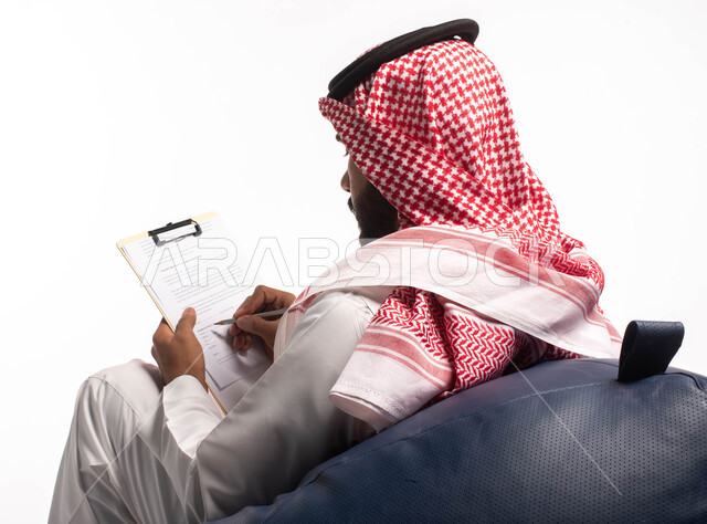 كتابة وتدوين الملاحظات الهامة، كتابة التقارير والابحاث على الورق، بورتريه من الخلف لشاب عربي خليجي سعودي يجلس على كرسي مريح ويحمل بيده أوراق وملفات خاصة بالعمل، خلفية بيضاء