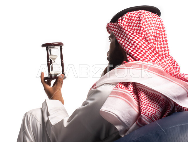 مراقبة وقياس الوقت, مفهوم إدارة الأعمال والوقت، بورتريه مقرب من الخلف لرجل سعودي خليجي يرتدي الثوب التقليدي يحمل بيده الساعة الرملية الزجاجية (البنكام)، توقيت الرمل، تحديد مواعيد العمل, إدارة الجدول الزمني، خلفية بيضاء. 