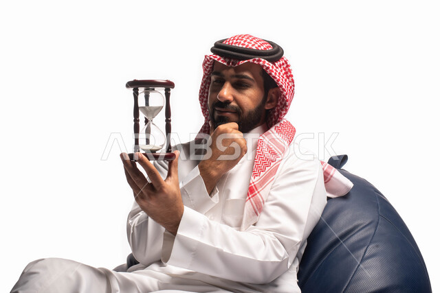 مفهوم إدارة الأعمال والوقت، بورتريه مقرب لرجل سعودي خليجي يرتدي الثوب التقليدي يحمل بيده الساعة الرملية الزجاجية (البنكام)، مراقبة وقياس الوقت, توقيت الرمل، تحديد مواعيد العمل, إدارة الجدول الزمني، خلفية بيضاء. 