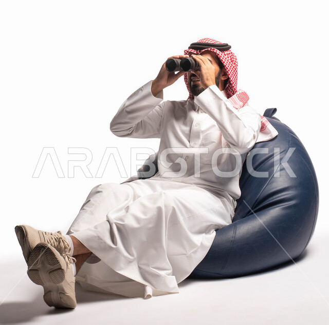 حب الاستكشاف والمغامرة، رؤية الاشياء البعيدة عبر المنظار، بورتريه لرجل عربي خليجي سعودي يرتدي الزي التقليدي يجلس على كرسي مريح وينظر من خلال منظار الرؤية, التخطيط المستقبلي للأهداف و الطموحات، خلفية بيضاء