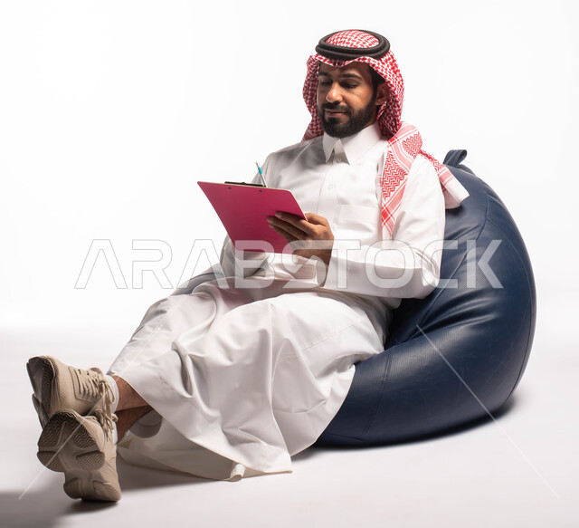 كتابة وتسجيل الملاحظات ، التمعن والتركيز ، إيماءات وجه ويدين تدل على التفكير ، بورتريه  لرجل عربي سعودي خليجي يرتدي الزي التقليدي يجلس على كرسي مريح ويمسك بيديه ورقة، خلفيه بيضاء