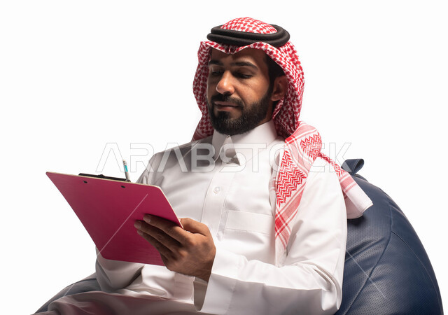 التمعن والتركيز ، كتابة وتسجيل الملاحظات ، إيماءات وجه ويدين تدل على التفكير ، بورتريه مقرب  لرجل عربي سعودي خليجي يرتدي الزي التقليدي يجلس على كرسي مريح ويمسك بيديه ورقة، خلفيه بيضاء