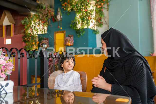 A Saudi Arabian Gulf child spends his time with his mother in the restaurant with gestures and facial expressions that indicate happiness, spending the holiday with the family, a family entertainment atmosphere, unique and distinctive sessions, restaurant
