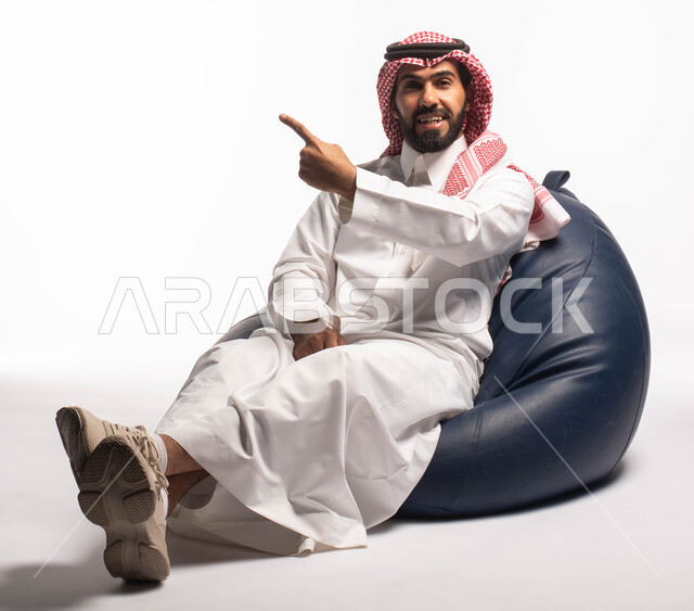 حركات يدين تدل على الإشارة إلى شيء ما, النظر إلى الكاميرا ، بورتريه لرجل عربي سعودي خليجي مبتسم يرتدي الثوب التقليدي يجلس على كرسي مريح, إيماءات وجه تدل على السعادة، خلفيه بيضاء