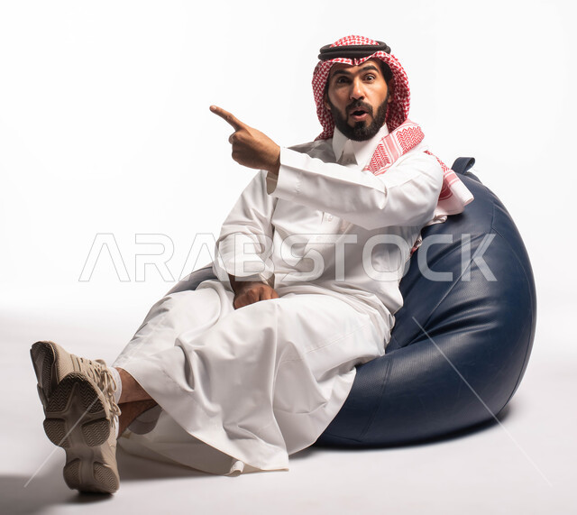 حركات يدين تدل على الإشارة إلى شيء ما, النظر إلى الكاميرا ، بورتريه لرجل عربي سعودي خليجي يرتدي الثوب التقليدي يجلس على كرسي مريح, إيماءات وجه تدل على الدهشة, خلفيه بيضاء
